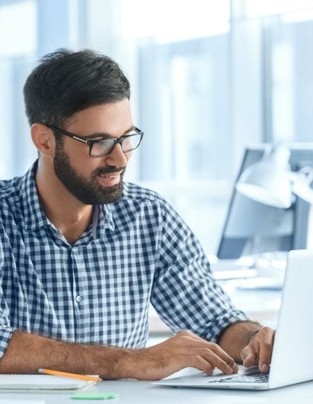 Mann mit Vollbart und Brille trägt ein kariertes Hemd und sitzt in einem modernen Büro vor einem geöffneten Laptop.